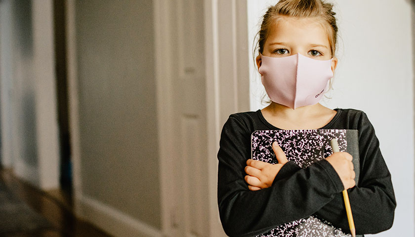 Blonde child wearing hair up, holding journal and wearing a mask