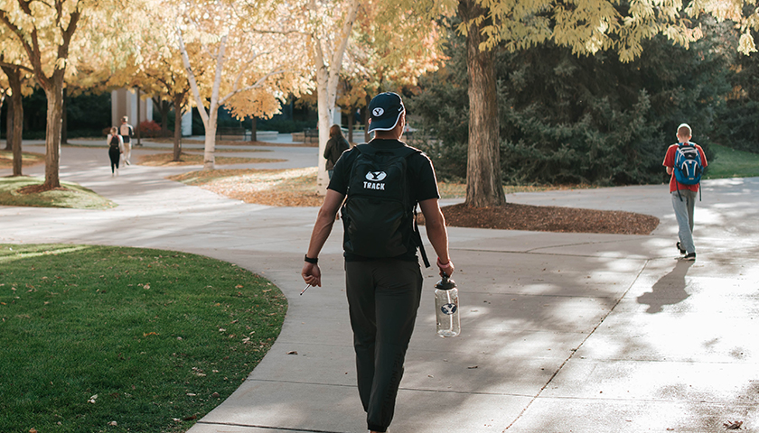 A man with a track backpack walks across the BYU campus in autumn.